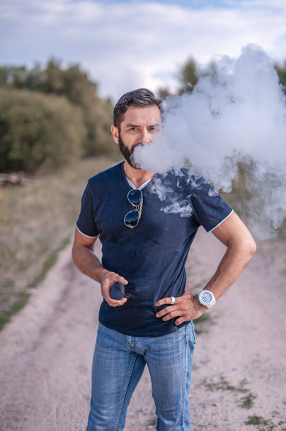Hombre de vapor barbudo disfrutando de un dispositivo de humo electrónico en la calma al aire libre. El cigarrillo de nueva tecnología. - Foto, Imagen