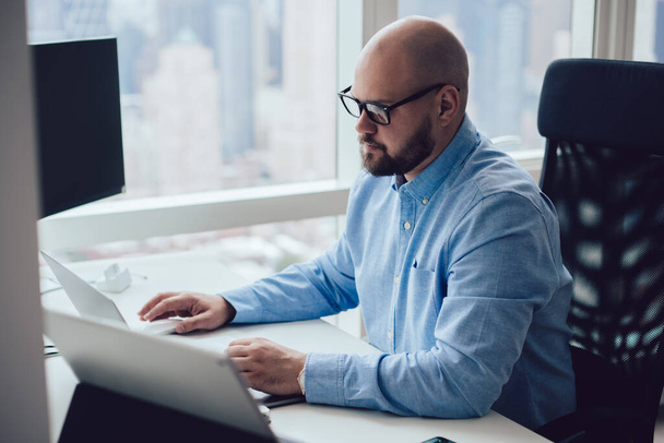 Concentrated bearded male employee wearing formal shirt and eyeglasses typing on keyboard of laptop while sitting in office near window and working hard - 写真・画像