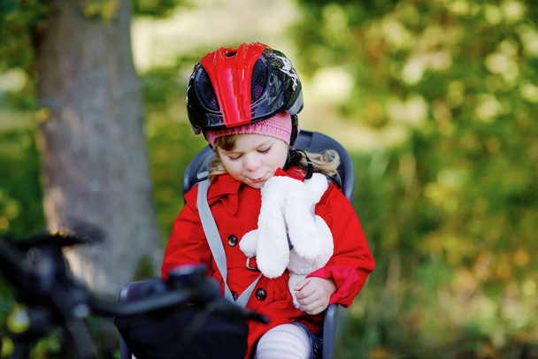 彼女の母親や父の自転車の自転車の座席に座って頭の上にセキュリティヘルメットを持つ小さな幼児の女の子.安全で子供の保護の概念。秋の日に家族旅行。子供保持プッシュソフトおもちゃ - 写真・画像