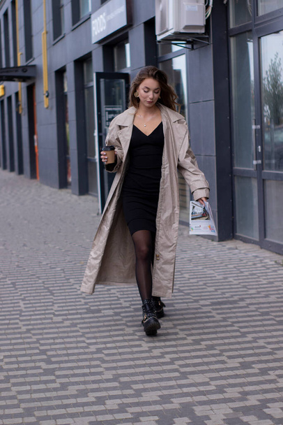 hermosa joven morena en gabardina de moda y vestido negro caminando por la calle. otoño, hojas amarillas. - Foto, imagen