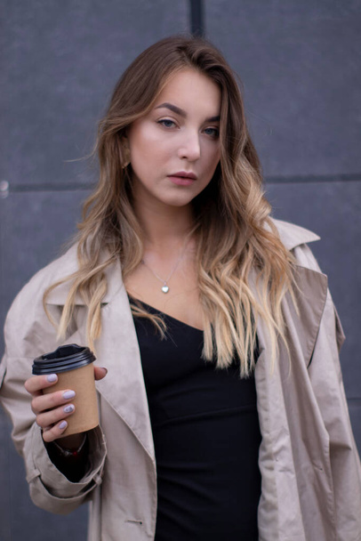 krásná štíhlá mladá brunetka v módní trenč kabát a černé šaty s papírovým šálkem kávy - Fotografie, Obrázek