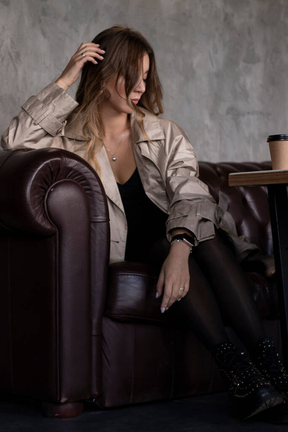 attractive slim brunette in trench coat in grey loft room on leather couch with paper cup. urban lifestyle concept - Photo, Image