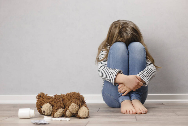 Triste adolescente, colegiala en depresión se sienta en el suelo cerca de la pared, junto a un oso de peluche y pastillas. - Foto, imagen