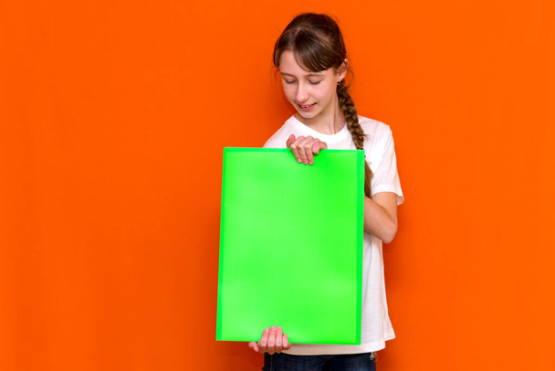Calm caucasian young girl holding a bright green folder with a banner advertisement on a orange studio background. advertising copy space - 写真・画像