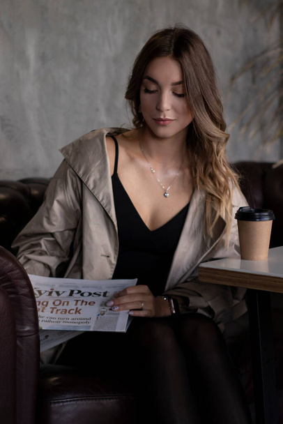jolie brune mince en trench coat dans la chambre grise loft sur canapé en cuir avec tasse en papier et journal. mode de vie urbain - Photo, image