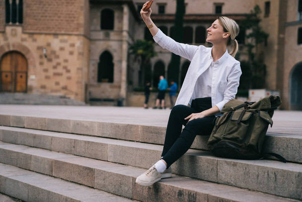 Full body of young female traveler sitting in city center with backpack and taking selfie near old buildings while spending weekend in Spain - Fotografie, Obrázek