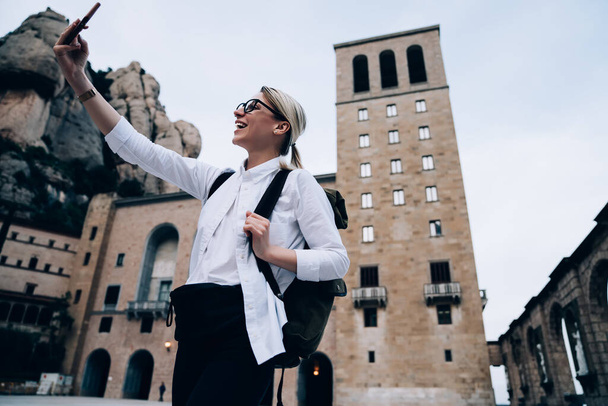 From below of excited smiling female tourist dressed in casual trendy outfit exploring Spanish sightseeing and taking picture of aged building in Montserrat mountains - Fotoğraf, Görsel