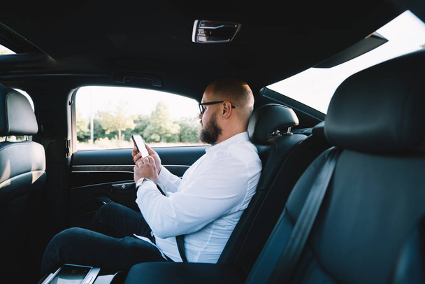 Side view of male worker with beard sitting in passenger seat in automobile and surfing mobile phone during ride to work - Foto, Bild