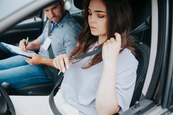 Attractive young woman nervous before driving test, fastening seat belt and looking down. Male auto instructor evaluating female student. Driving lessons. Obtaining a drivers license concept. - 写真・画像