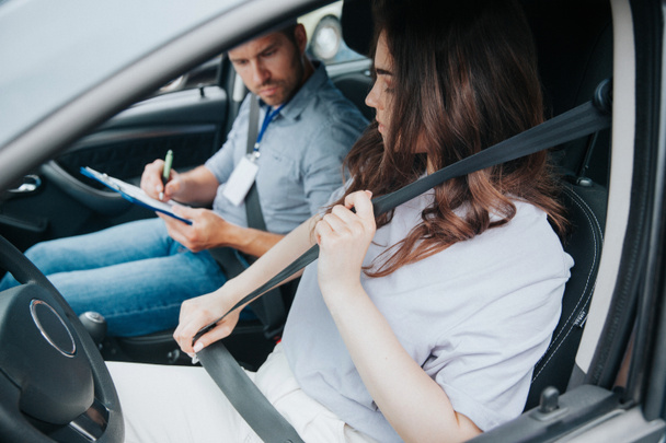 Serious young woman fastens her belt. Concentrated driving instructor makes notes on his clipboard. Beautiful young woman with wavy brown hair in white clothes takes driving lessons. - Фото, изображение