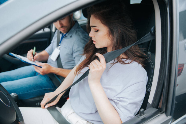 Beautiful young woman upset with results of driving tests. Strict examiner holds clipboard and makes notes of her mistakes. Female student and male instructor sit in a car with open window. - Fotografie, Obrázek