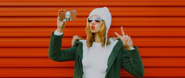 young woman taking selfie picture by phone wearing a knitted hat, sweater on red background - Foto, afbeelding