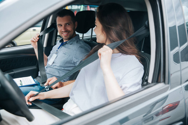 Young female student and attractive male driving instructor smile to each other sitting in the car and fastening seat belts. Positive experience with driver courses concept. - Photo, image