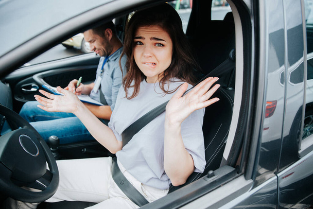 Stressed young woman on drivers seat of black car throws up her hands in confusion and bites her lip. Upset driving examiner makes notes in clipboard. Problems on driving test concept. - Foto, Imagem