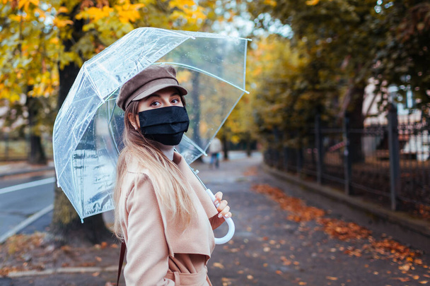 Portrait of young woman in protective mask walking along autumn city street under transparent umbrella during rain. Coronavirus covid-19 pandemic - Photo, Image