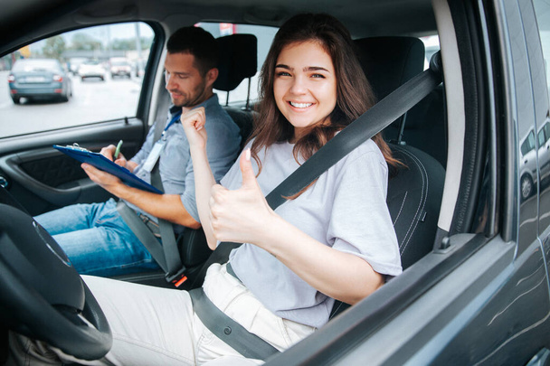 Szczęśliwa młoda kobieta podnosi kciuk. Uczennica uśmiecha się z powodu pomyślnego egzaminu na prawo jazdy, patrzy w kamerę i wskazuje na instruktora jazdy. - Zdjęcie, obraz