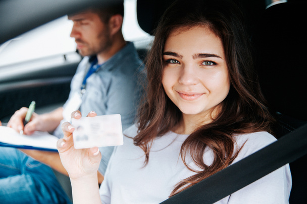 Ελκυστική νεαρή γυναίκα που δείχνει άδεια οδήγησης, κοιτάζει την κάμερα και χαμογελά. Μελαχρινή γυναίκα κάθεται στο κάθισμα του οδηγού με δεμένη ζώνη ασφαλείας. Θολή αρσενικό εκπαιδευτή στο παρασκήνιο γράφοντας κάτι. - Φωτογραφία, εικόνα