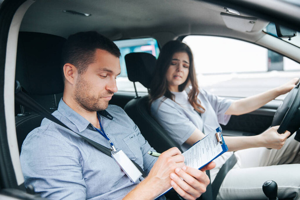 Upset woman failed driving exam bites lips and worries about it. Male instructor notice results of test in his paper. Female student holding wheel and looking troubled at the driving instructor. - Φωτογραφία, εικόνα