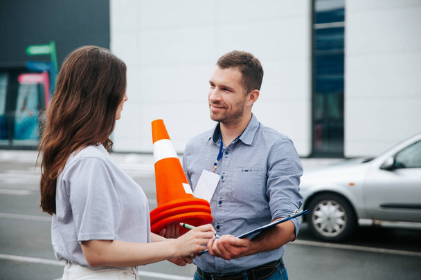Female student helps to pack traffic cones, male driving instructor smiles with gratitude. Young woman in white clothes and attractive man in gray shirt together hold orange traffic cones. - Photo, Image
