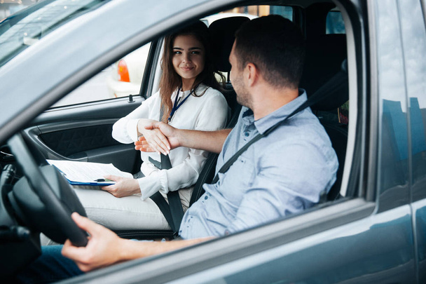 Female auto instructor shaking hands with male student. Attractive woman congratulates with successful driving license exam. Man in blue shirt holding wheel by his hand and looking at woman. - Photo, Image