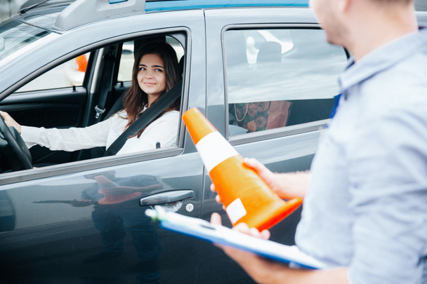 Die attraktive brünette Frau auf dem Fahrersitz mit angeschnalltem Sicherheitsgurt lächelt den Fahrlehrer mit Klemmbrett und orangefarbenem Verkehrskegel in den Händen an. Konzept zur Verbesserung der Fahrkompetenz. - Foto, Bild