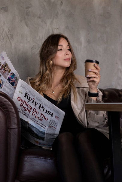 attractive slim brunette in trench coat in grey loft room on leather couch with paper cup and newspaper. urban lifestyle - Foto, Imagem