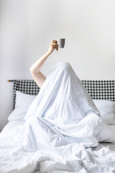 Молода жінка з чашкою кави або чаю сидить у ліжку під ковдрою у спальні. Перебування вдома
 - Фото, зображення