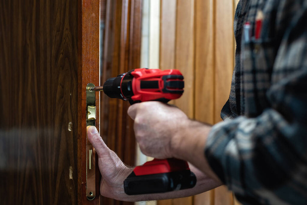 Close-up. O carpinteiro com uma chave de fenda elétrica fixa a fechadura de uma porta de madeira. Indústria da construção.  - Foto, Imagem