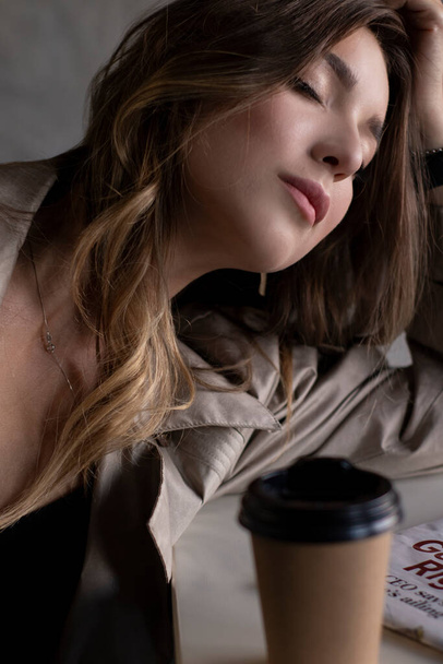 atrakcyjna szczupła brunetka w płaszczu w szarym pokoju na poddaszu na skórzanej kanapie z papierową filiżanką i gazetą. miejski styl życia - Zdjęcie, obraz