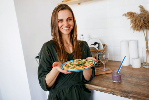 Молодая девушка с широкой улыбкой на кухне держит вегетарианскую пиццу. - Фото, изображение