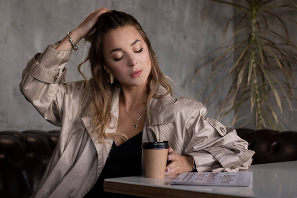 atraktivní štíhlá brunetka v plášti v šedém podkrovním pokoji na koženém gauči s papírovým kelímkem a novinami. městský životní styl - Fotografie, Obrázek