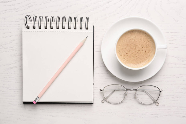 Notebook with pencil, glasses and cup of coffee on white wooden table, flat lay - Фото, изображение