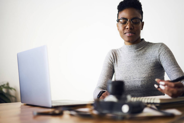 Positive black female freelancer wearing eyeglasses and casual sweater taking notes in notepad while sitting at table with modern laptop - Fotó, kép