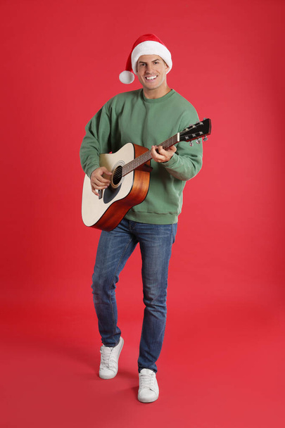 Man in Santa hat playing acoustic guitar on red background. Christmas music - Φωτογραφία, εικόνα