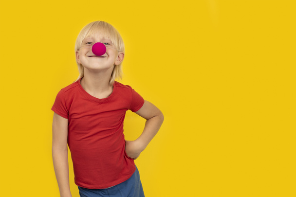 Niño feliz vistiendo la nariz de payaso y sonriendo a la cámara. Retrato de niño sobre fondo amarillo. - Foto, Imagen