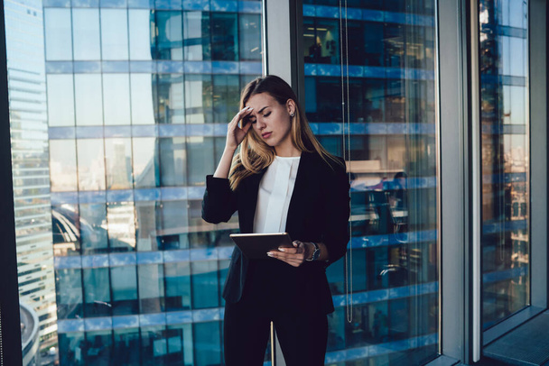 Exhausted young female entrepreneur in formal suit standing with closed eyes near window in contemporary glass building and browsing tablet while frowning and touching head - Foto, immagini