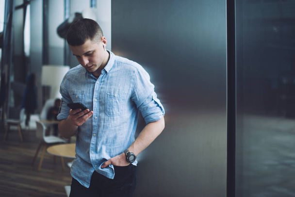 Focused man in casual clothes and wrist watch and wireless earphones standing near metal wall against cafe furniture while browsing Internet on mobile phone - Photo, Image