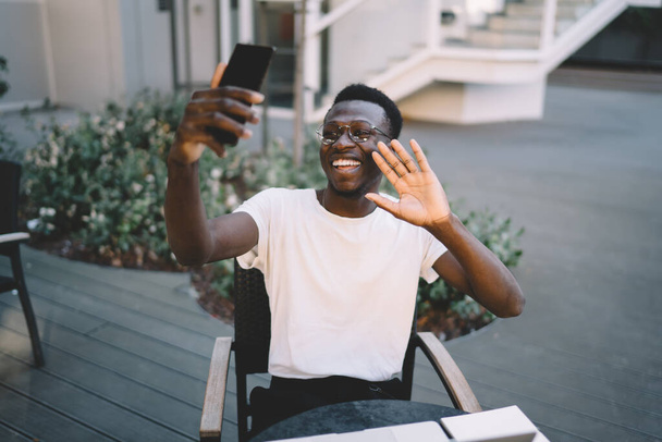 Cheerful dark skinned african american male blogger using smartphone camera for making vlog video sitting on cafe terrace, cheerful african american man waving during mobile phone online call - Foto, afbeelding