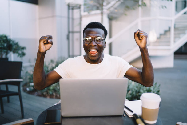 Πορτρέτο του χαρούμενου σκουρόχρωμου άνδρα blogger στο eyewear γιορτάζει ευημερούσα έργο σε απευθείας σύνδεση εργάζονται εξ αποστάσεως στην καφετέρια βεράντα, χαμογελώντας νεαρός Αφροαμερικανός απολαμβάνοντας τη νίκη σε video game - Φωτογραφία, εικόνα