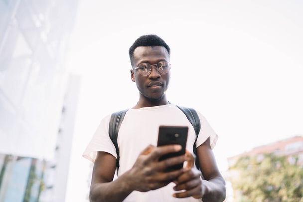 Pensive african american male blogger share multimedia content online standing with mobile phone in city, pensive dark skinned hipster guy in casual wear and eyeglasses dialing number on smartphone - Φωτογραφία, εικόνα