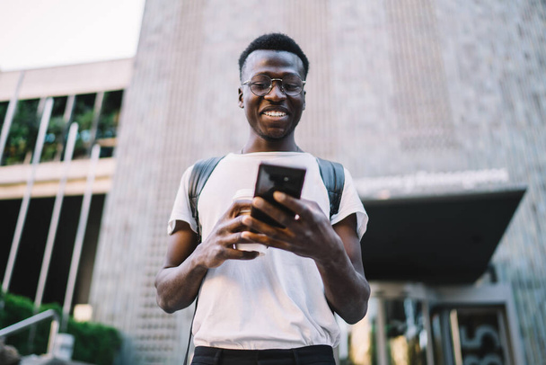 Cheerful african american hipster guy in trendy casual wear strolling with smartphone on city street, young dark skinned male millennial blogger sending text messages online using mobile phone - Foto, afbeelding