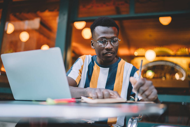 Serious black male student in striped tshirt and glasses taking notes while sitting at table with laptop and working on project in cozy cafeteria - 写真・画像