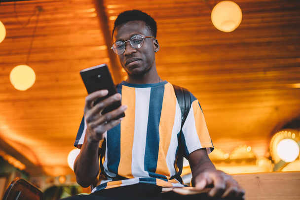 From below of serious young African American guy in casual wear and eyeglasses browsing smartphone while resting in cafeteria with wooden interior and waiting for order - Fotografie, Obrázek