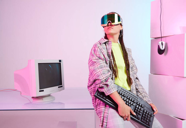Stylish young female in goggles with old computer monitor, mouse, keyboard on workplace in office. Colored Neon light - Foto, afbeelding