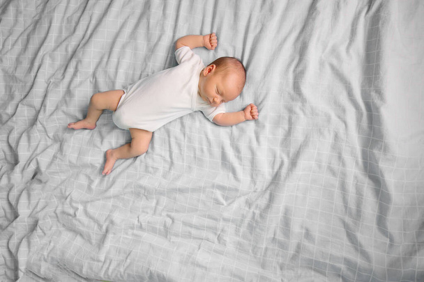 The tiny newborn is sleeping on the parent's bed. Place for text. Innocent Infant Boy in white bodysuit. Gray bedroom background. Childhood parenting. Sweet dream.Real life lifestyle small moments - Foto, Imagen
