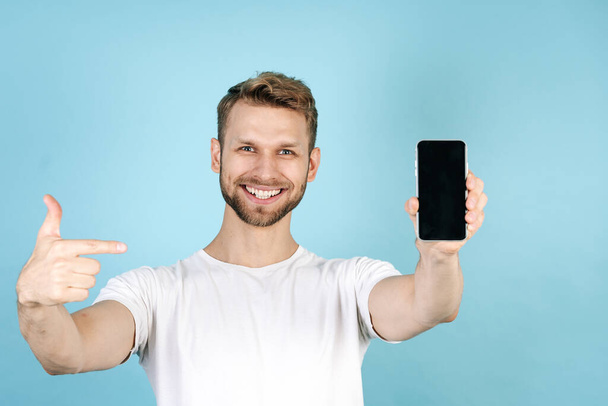 Concept of sale and discount in online store. Smiling and happy young man pointing at modern smartphone with blank screen display, standing isolated on blue copy space background in white t-shirt - Photo, image