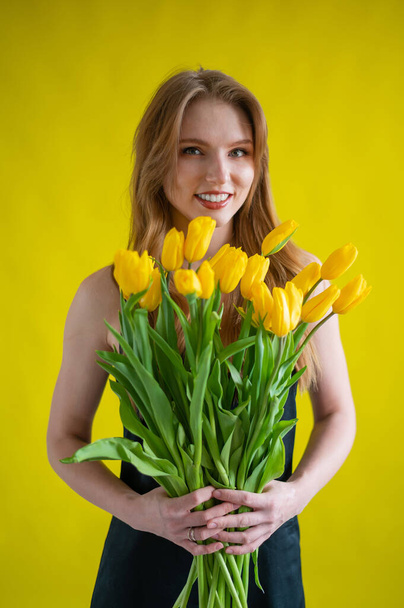 Kaukasische Frau mit einem Arm voller gelber Tulpen auf gelbem Hintergrund. Internationaler Frauentag. Strauß Frühlingsblumen - Foto, Bild
