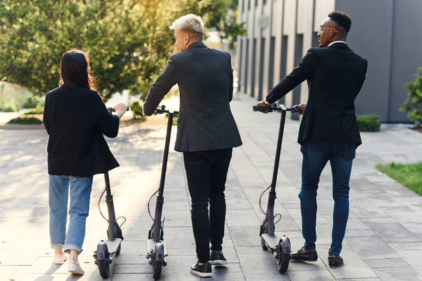 Professional multi ethnic co workers hold electric scooters walking near modern building and talking - Valokuva, kuva