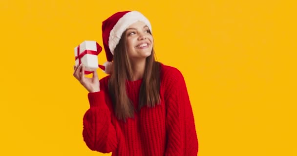 Noel Baba şapkalı genç ve meraklı bir kadın Noel hediyesini sallıyor, hediye kutusundaki tıkırtıları dinliyor. - Video, Çekim
