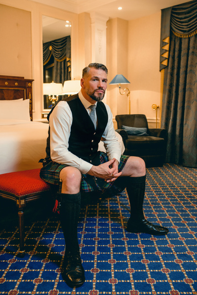 guapo maduro valiente hombre elegante escocés en kilt en habitación de hotel de lujo. Estilo, moda, estilo de vida, cultura, viajes, concepto étnico. - Foto, Imagen